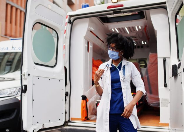 African american female paramedic in face protective medical mask standing in front of ambulance car.